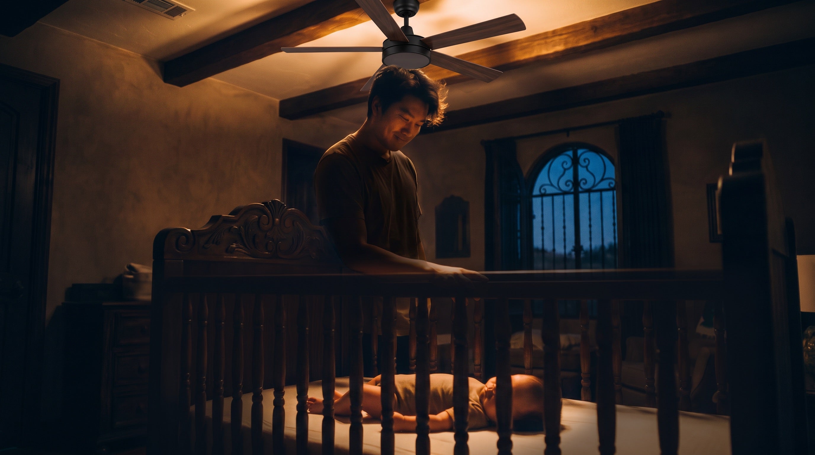 A father beside his baby’s crib in a nursery beneath a ceiling fan, the baby resting on its back.